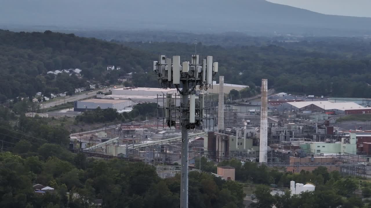 Drone orbit of cell tower overlooking an industrial complex in America, with factories, warehouses and power infrastructure surrounded by forest and mountains in distance. Cloudy sky in waynesboro,usa