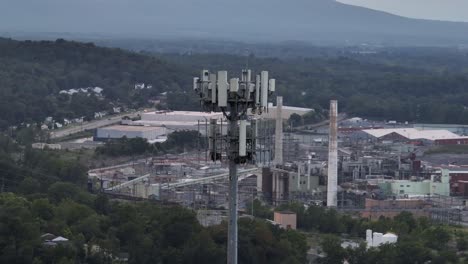 Drone orbit of cell tower overlooking an industrial complex in America, with factories, warehouses and power infrastructure surrounded by forest and mountains in distance. Cloudy sky in waynesboro,usa