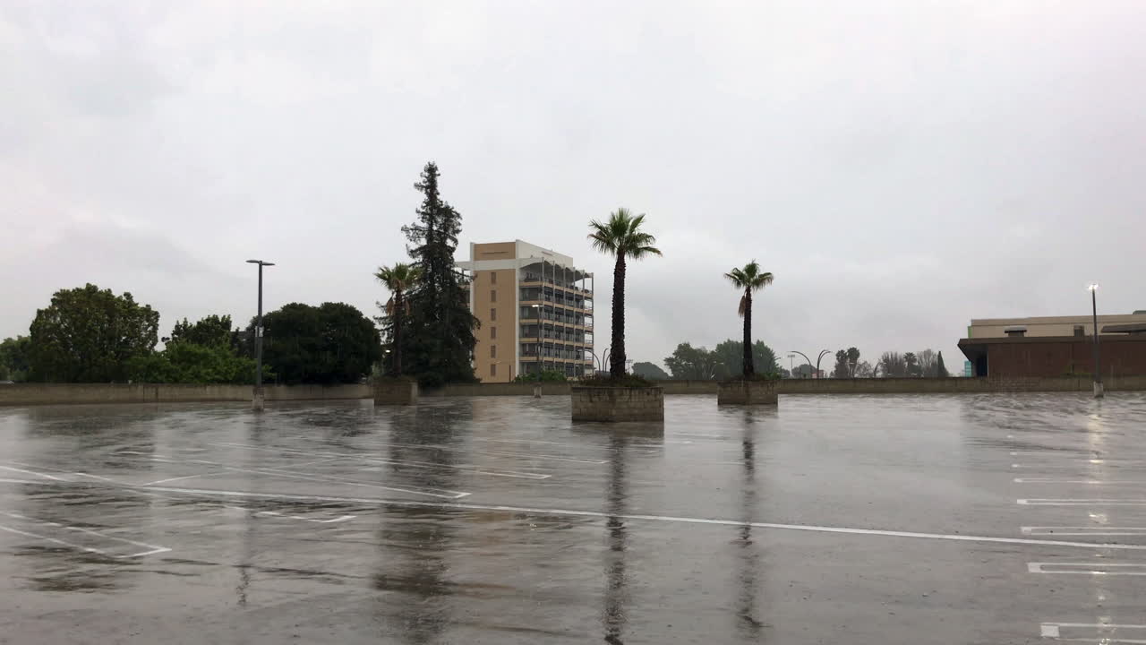 la lluvia cae en un estacionamiento vacío en un día oscuro con palmeras y edificios a distancia