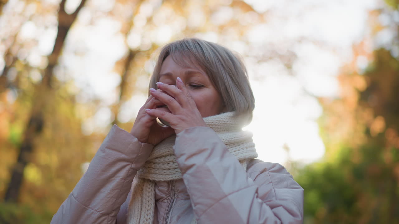close up of woman holding cup of tea near lips, gently blowing to cool steaming drink before sipping, wrapped in chunky knit scarf against blurred autumn foliage and warm sunlight glow