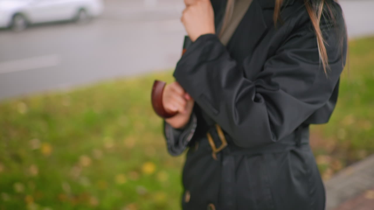 도시 거리에서 검은색 코트를 입은 사람이 나무 곡선 손잡이가 달린 검은색 닫힌 우산을 들고 서 있는 클로즈업 사진이에요. 이는 기대, 준비, 날씨 보호, 회복력, 개인 안전을 상징합니다.