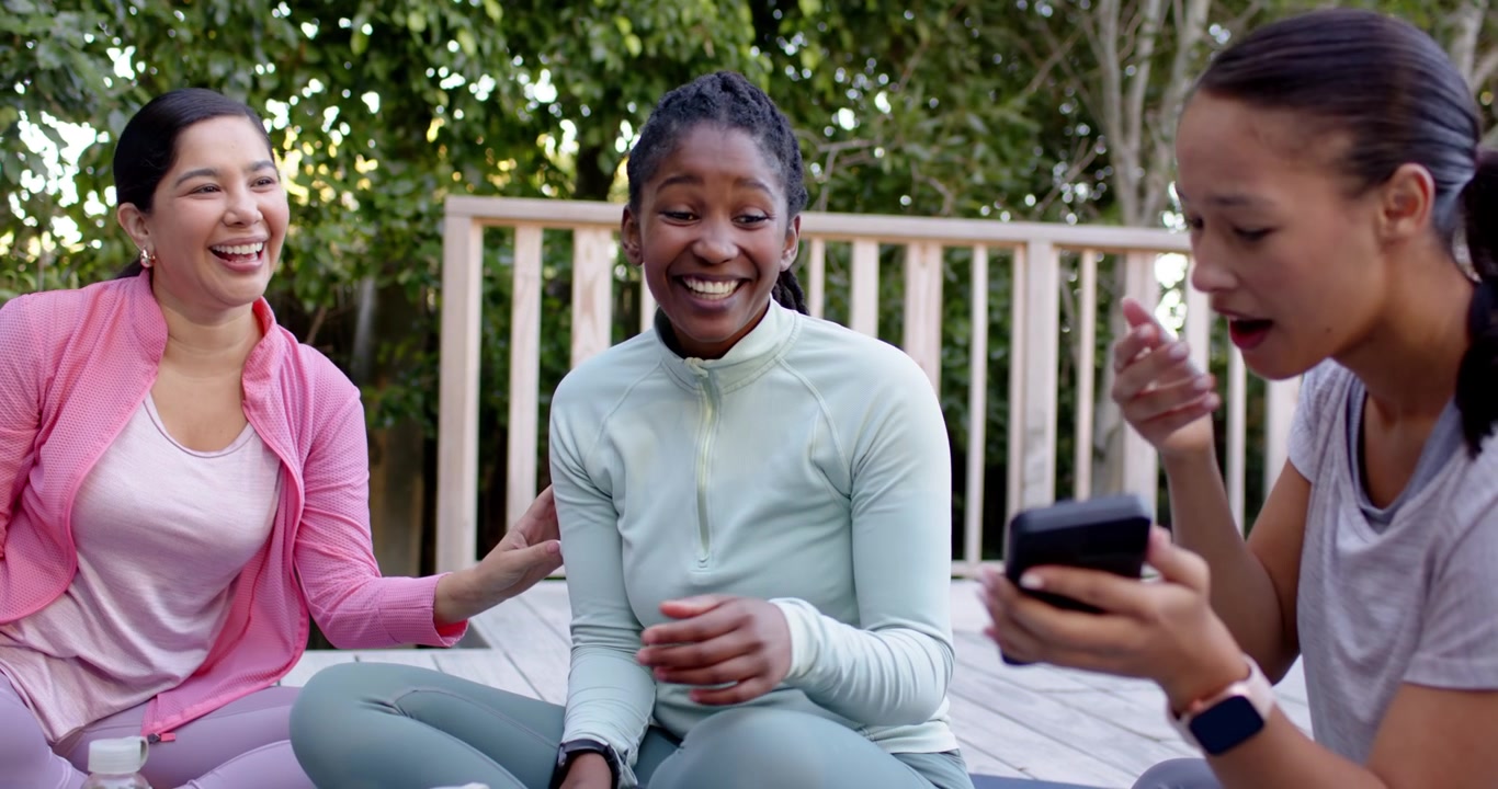 Diverse female friends laughing on exercise mats on deck as Asian woman holding phone showing joke