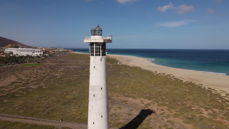 toma aerea en orbita ya media distancia del faro de morro jable, con los edificios de la ciudad y la playa de morro jable al fondo