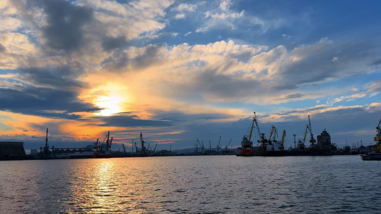 Sunset over harbor with cranes and water reflecting light. The sky showcases colors as the sun sets behind cranes by the water. Boats are stationary as clouds drift above