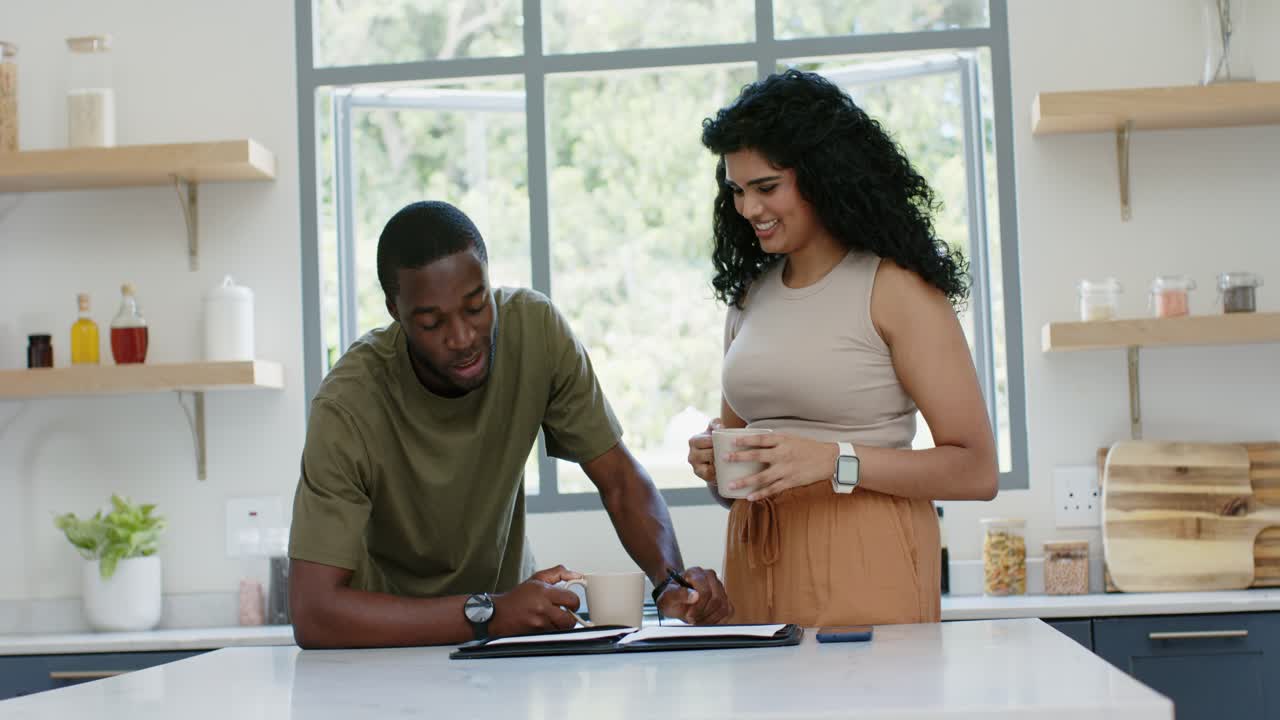 Diverse couple, man tapping tablet in kitchen, woman stepping closer, reviewing content with mugs