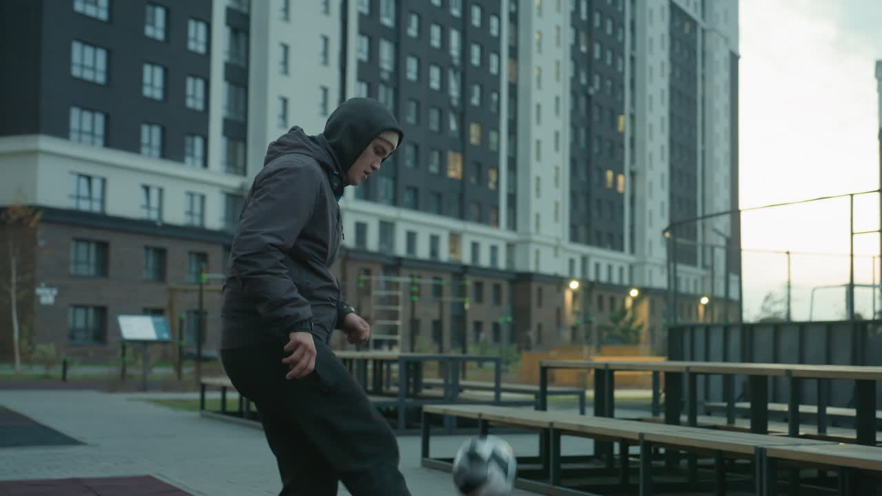 Athlete skillfully juggling football on sport arena with urban background featuring benches, lit pathways, and tall residential buildings