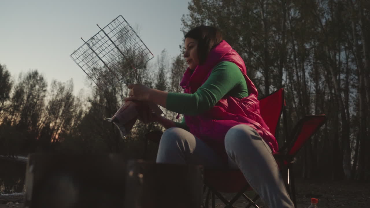 Donna caucasica che si occupa del fuoco al tramonto in campeggio, seduta su una sedia pieghevole, che regola la griglia metallica sopra le braci ardenti, con gilet rosa e maglione verde, sagome di alberi al tramonto, calma, concentrata, da sola.