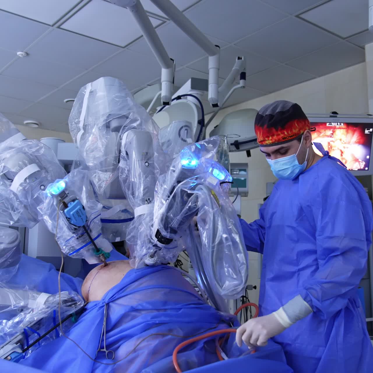 Caucasian surgeon in bright cap and mask stands near the robotic equipment in modern surgery room. Screen at backdrop showing the operational procedure
