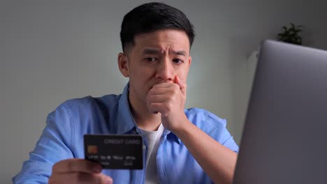 serious young asian man, frowning and worried while using and looking at credit card, doing online shopping, making online payment transaction using laptop at home.