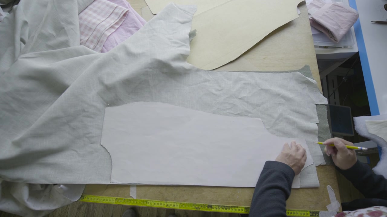 Shot from above showing a hand drawing and marking with a yellow pencil the outlines of a pattern piece on a spread white fabric