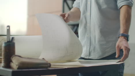 Hipster engineer works with blueprint. Shoot from above. Young architectural engineer working in office. Bearded man makes sketches. Loft style minimalistic interior drawings on the table