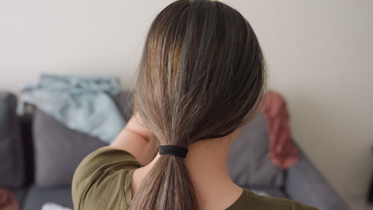 Close up rear view of lady with tied ponytail wearing green shirt placing hand on head showing fatigue while sitting on couch with scattered clothes in messy room environment