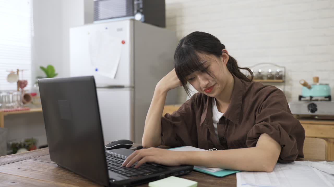 편두통을 고 있는 아픈 아시아 젊은 여성은 불편함으로 머리를 고 식탁에서 집에서 일하면서 컴퓨터에서 타이핑을 계속합니다.
