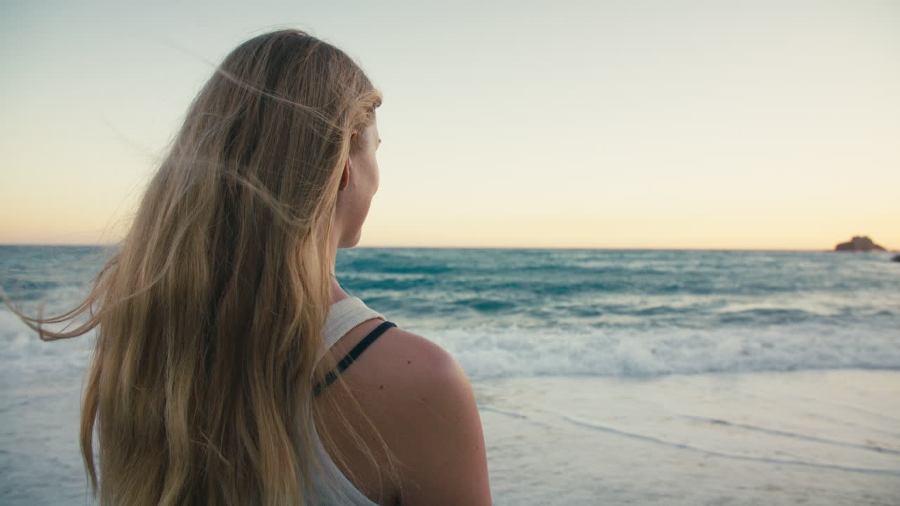 Rear medium close-up of a woman with flowing blonde hair as she gazes at the sea, her hair gently lifted by the wind at sunset