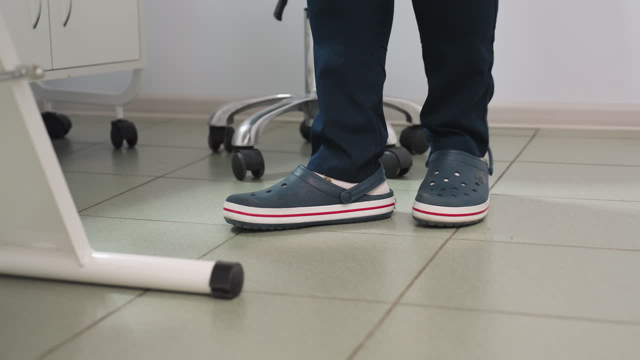 Leg view of clinician in crocs standing from seat and moving to side of massage chair in clean spa environment with tiled floor and equipment trolley visible wearing navy uniform lamp near station