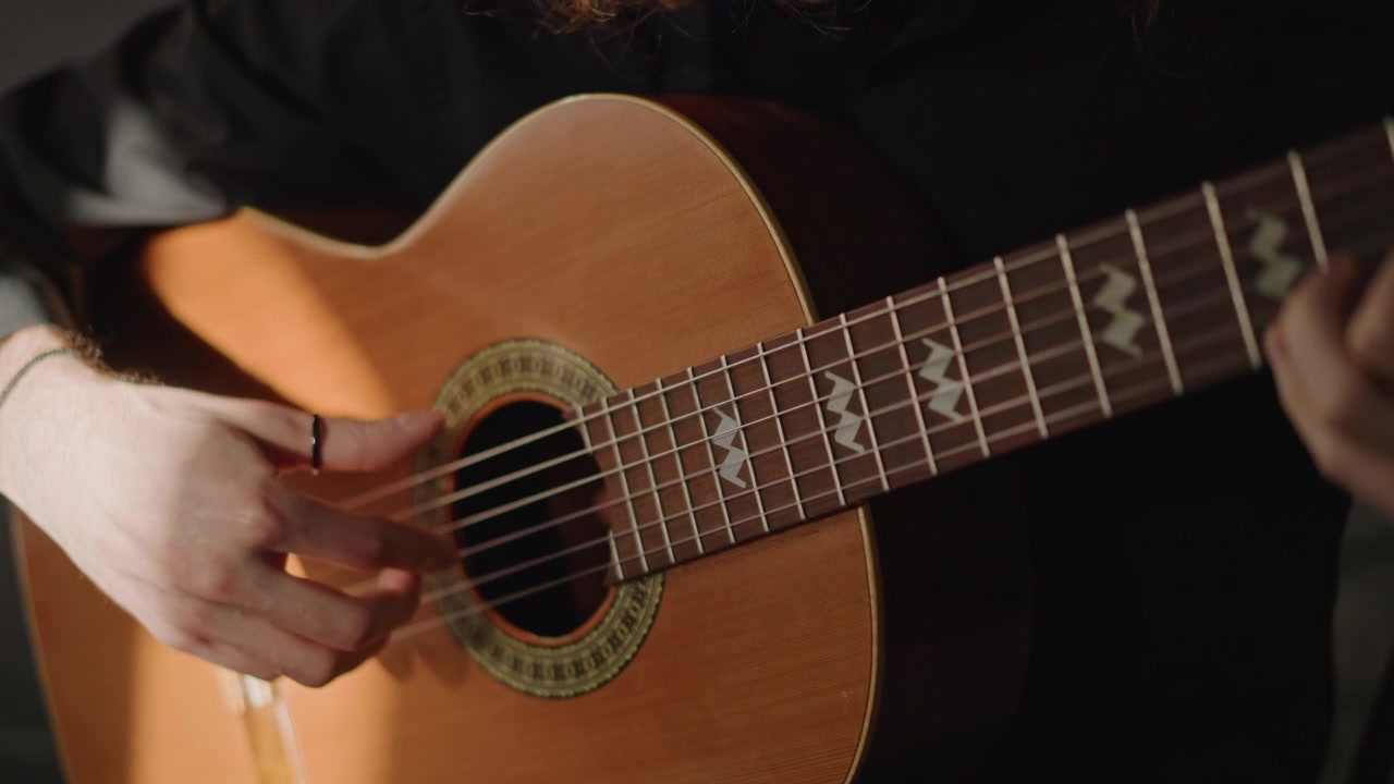 Primer plano de guitarra acústica, mano pulsando cuerdas de nailon en estudio de poca luz, ejercicio de fingerpicking concentrado por guitarrista masculino, tonos de madera cálidos y sombras suaves que transmiten concentración y maestría.