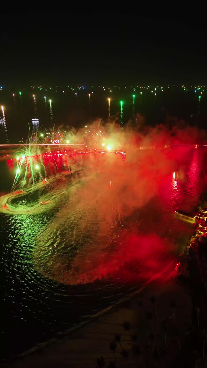 Vertical aerial night view of a vibrant water and laser show at Sunset Town on Phu Quoc Island, Vietnam, with colorful lights reflecting off the water and surrounding buildings illuminated.