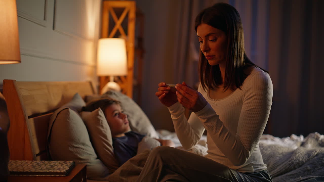 Primo piano di una mamma premurosa che controlla la temperatura del figlioletto sdraiato comodamente nel letto di casa.