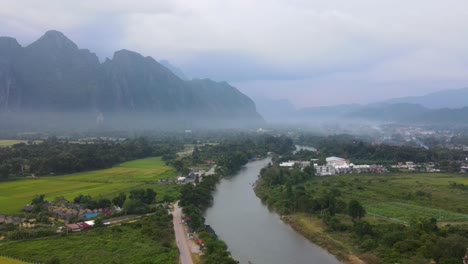 Luftaufnahme-Von-Vang-Vieng-Am-Abend-Mit-Dem-Fluss-Nam-Xong,-Der-Durch-Eine-Neblige-Landschaft-Fließt