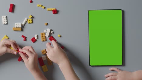 overhead shot of two children playing with plastic construction bricks next to green screen digital tablet