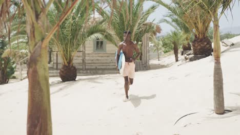 Happy-african-american-man-carrying-surfboard-running-on-sunny-beach,-slow-motion