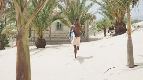 happy african american man carrying surfboard running on sunny beach, slow motion