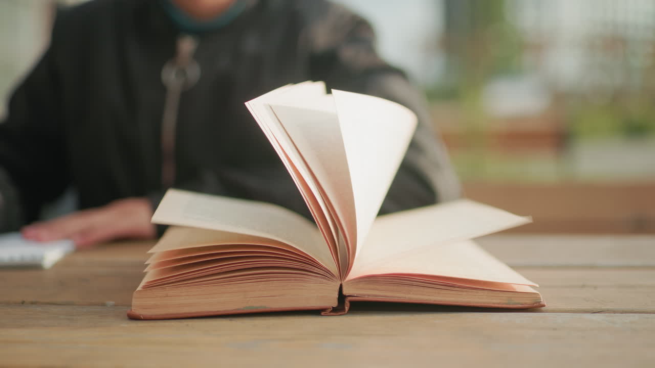 Close up of kid flipping to new page of open book on wooden table outdoors with blurred bokeh background, novel pages lifting slightly in breeze while kid writes in notebook