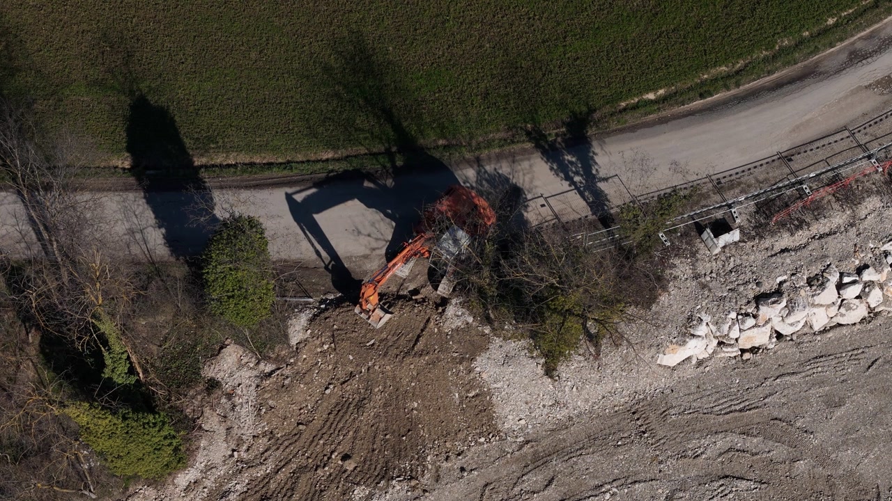 Hydraulic excavator performing earthworks along rural road, showing digging activity and construction site with soil movement and infrastructure work from aerial top view.