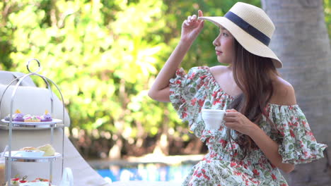 Schöne-Asiatische-Frau-In-Kleid-Und-Schlapphut,-Die-Im-Baumschatten-Mit-Einer-Tasse-Getränk-Am-Pool-Des-Luxuriösen-Tropischen-Resorts-Sitzt