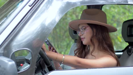 A woman in glasses and hat smiling while using her mobile phone in the driver's seat of a car