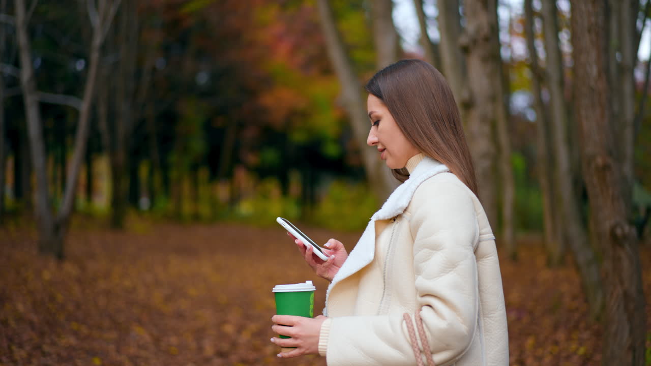 Romantic beautiful lady walking by the autumn park. Woman holding a paper cup in her hand and looking at smartphone. Blurred backdrop.