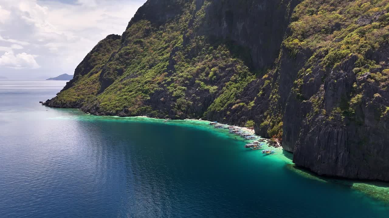 Outrigger boats line the clear turquoise waters of Talisay Beach, surrounded by steep limestone cliffs and dense greenery on Tapiutan Island, Philippines, in a peaceful tropical setting