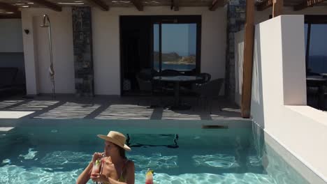 Brunette-woman-in-swimming-costume-is-in-a-pool-at-Cayo-Resort-in-Greece-Crete---she-enjoys-the-water-and-drinks-a-cocktail---drone-flies-up-and-you-can-see-the-resort