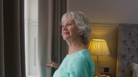 smiling senior woman at home wearing pyjamas opening bedroom curtains and looking out of window