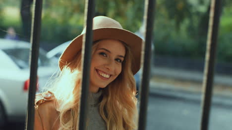 alegre joven rubia sonriente con un sombrero y un abrigo caminando detrás de una cerca mientras mira la cámara en la calle