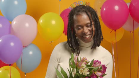 studio portrait of woman wearing birthday headband holding bunch of flowers celebrating with balloons 1