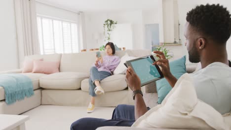 Diverse-couple-sitting-on-couch-and-using-tablet-in-living-room