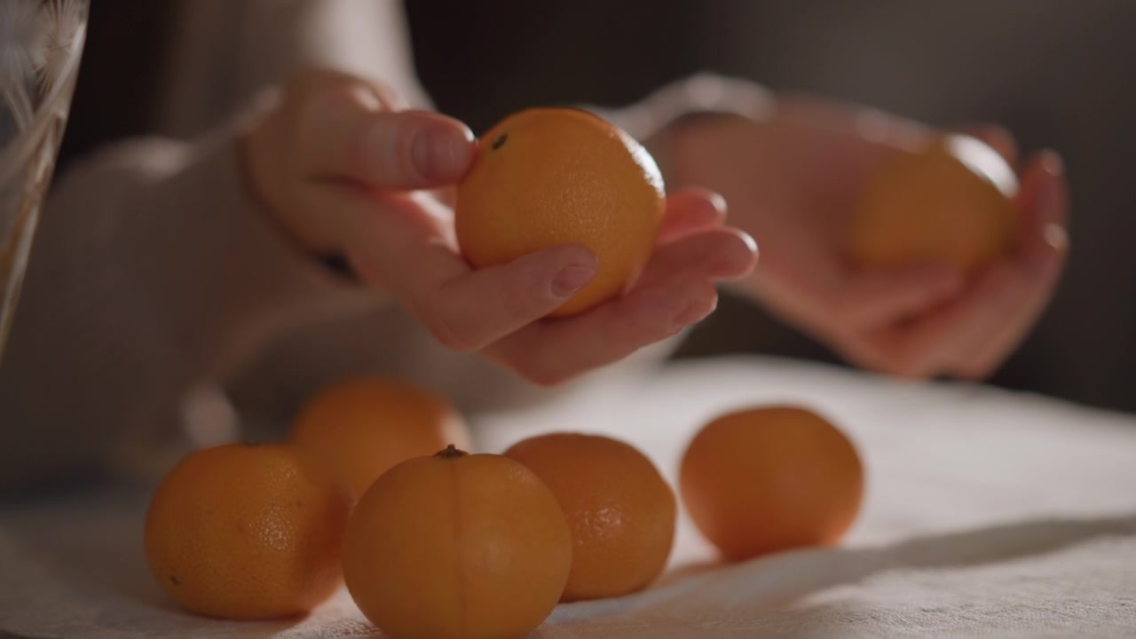 Close-up de mãos femininas caucasianas segurando clementinas, luz suave da janela, toalha de mesa de linho, classificação tátil lenta, contagem, preparação de lanche, casca texturizada, tons quentes e suaves, estudo de estilo de alimentos, doméstico.