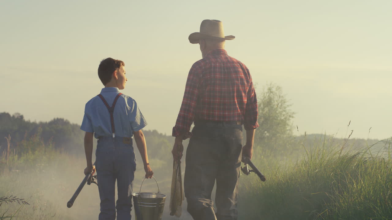 vista trasera del abuelo caucásico y su nieto caminando juntos mientras sostienen cañas de pescar por la mañana