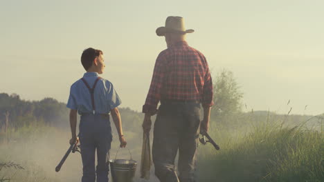 rear view of caucasian grandfather and his little grandson walking together while holding fishing rods in the morning