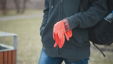 black guy wearing dark gray textured hoodie holding bright orange gloves in one hand, carrying black backpack with water bottle, standing outdoors in park during early spring, blurred grassy background