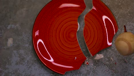 ceramic red plate with a spiral pattern being crushed to pieces by a baseball hitting it in slow motion on a concrete fool