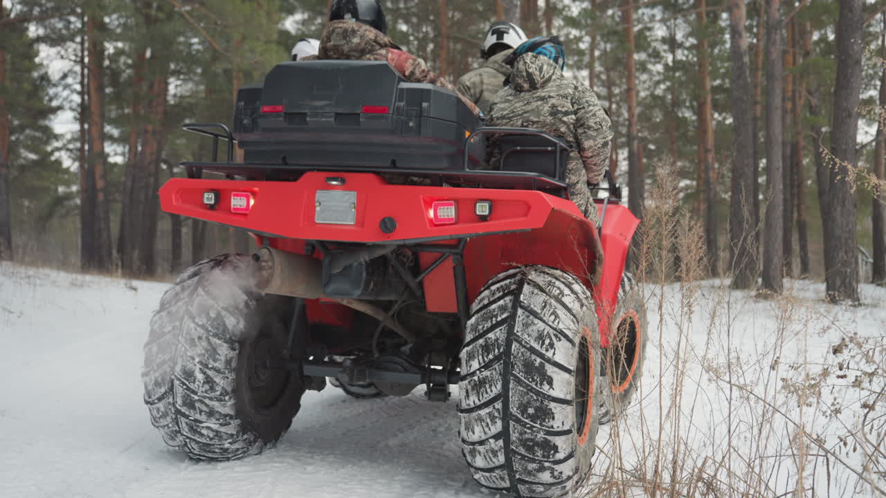 Pneumatici ATV in primo piano che sollevano la neve mentre un quad rosso sfreccia su un sentiero ghiacciato, con spruzzi dalle ruote posteriori e scarico visibile. Due piloti con caschi accelerano attraverso una pineta, in un'azione fuoristrada ad alta trazione.