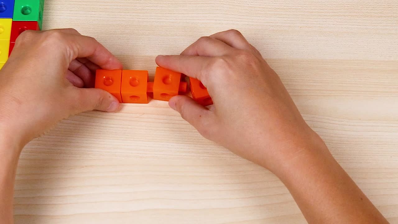 Close-up of hands assembling orange linking cubes on a light wooden surface.
