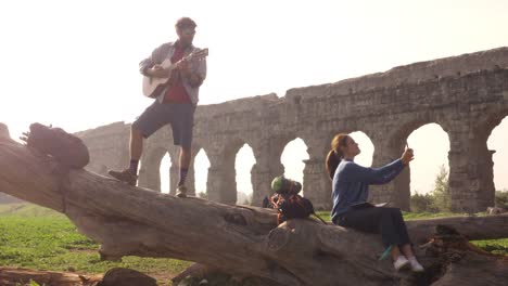 happy young couple backpackers tourists on a log trunk taking selfies photos with smartphone in front of ancient roman aqueduct ruins in romantic parco degli acquedotti park in rome misty sunrise slow motion