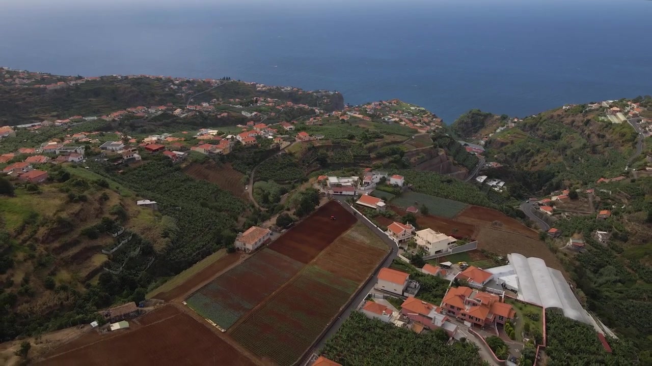 Ripresa con drone in movimento all'indietro che sorvola terrazzamenti agricoli e case rurali, con l'Oceano Atlantico che diventa più prominente man mano che la telecamera avanza verso l'orizzonte