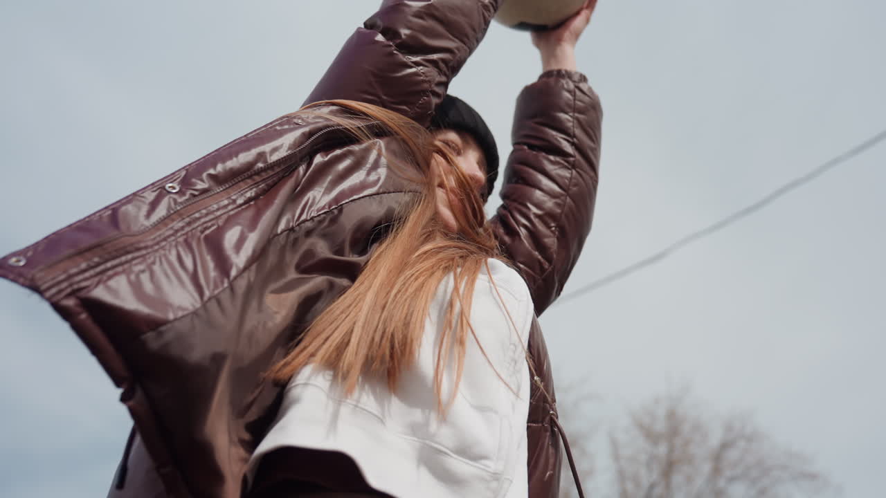 young white woman stretching arms skyward outdoors, low-angle confident pose in brown puffer jacket and black beanie, long hair flowing, overcast sky, city field backdrop, energetic candid portrait