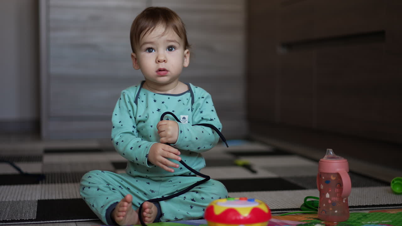 Little kid in blue pajama sitting on the floor holding a black lace in his hands. Lovely child playing on the floor at home. Blurred backdrop.