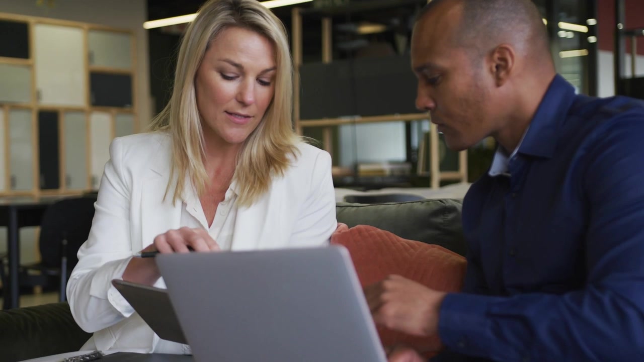 Woman tapping tablet directing man adjusting laptop for business review with bokeh overlays passing