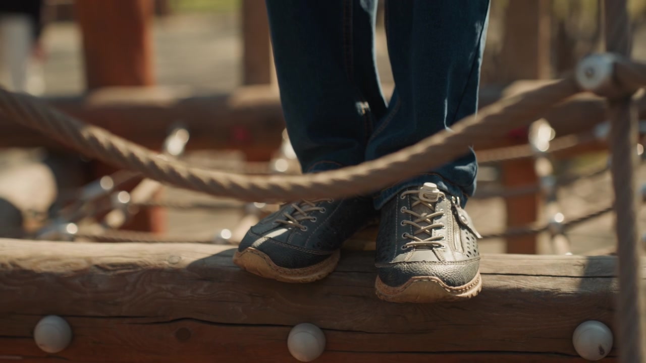 Nærbillede af barnefødder i blå jeans og sneakers, der forsigtigt træder over træstammer med reb, balancering på en udendørs forhindringsbane. Fokus forbliver udelukkende på underben og sko, mens de